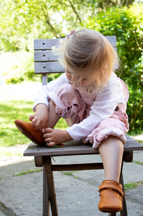 petite fille assise sur une chaise dehors en train d'enfiler des chaussons en cuir souple Achille marron à scratch