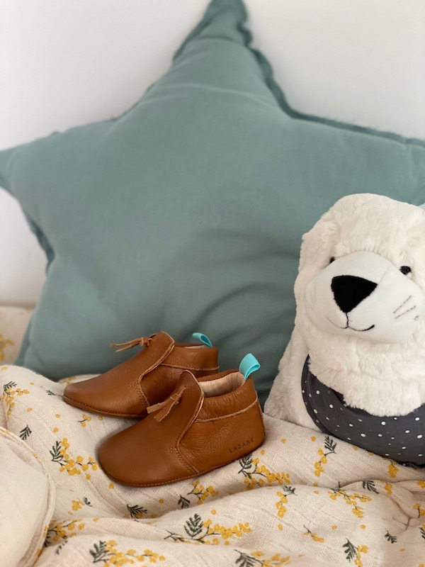 chaussons bébé en cuir souple marron Madeleine avec pompon sur le dessus posé sur un foulard avec coussin étoile en fond