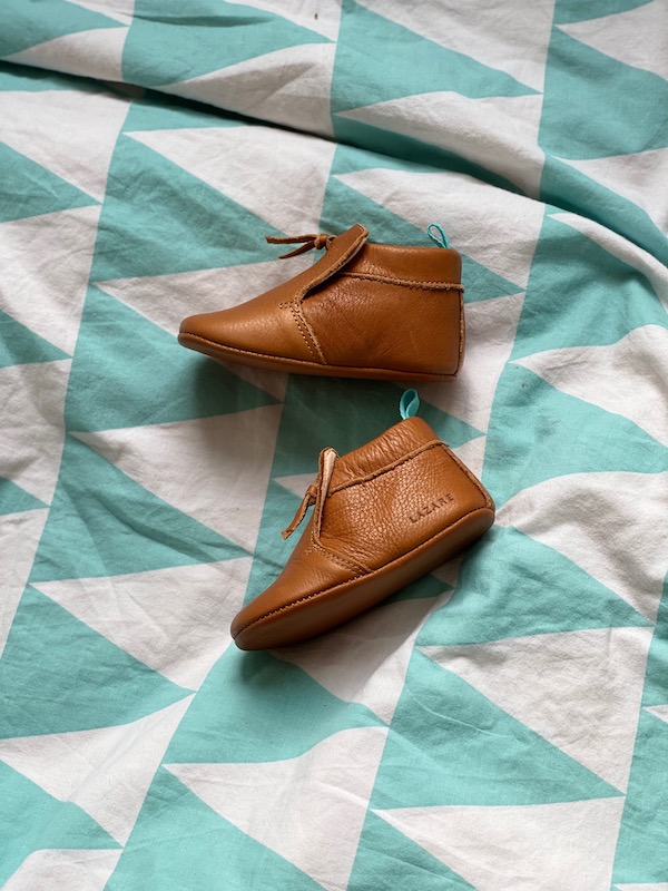 chaussons bébé en cuir souple marron Madeleine avec pompon sur le dessus posés sur un lit