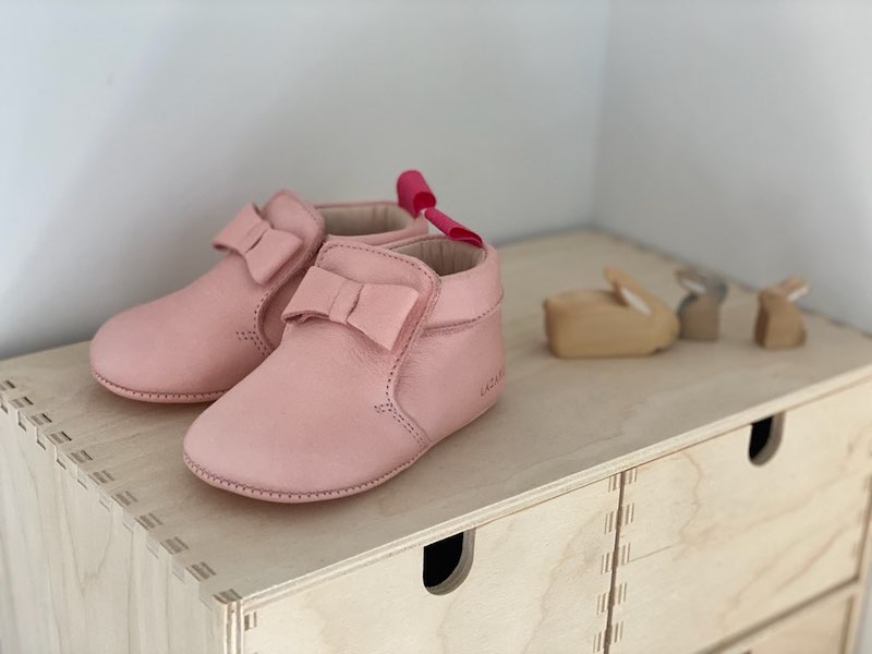 chaussons bébé Arielle en cuir souple rose avec noeud sur le dessus posés sur une étagère en bois