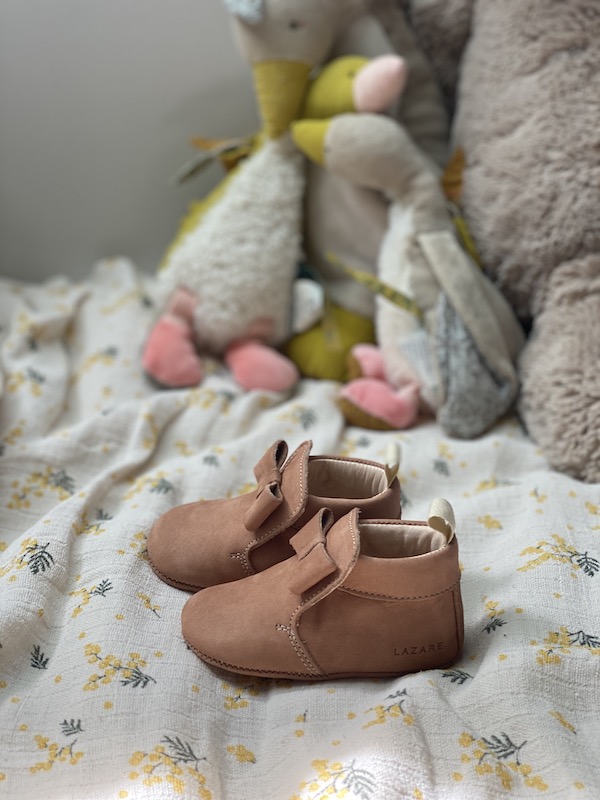 chaussons bébé Arielle en cuir nubuck rose souple avec noeud sur le dessus vu de coté posé sur un lit avec peluches en fond